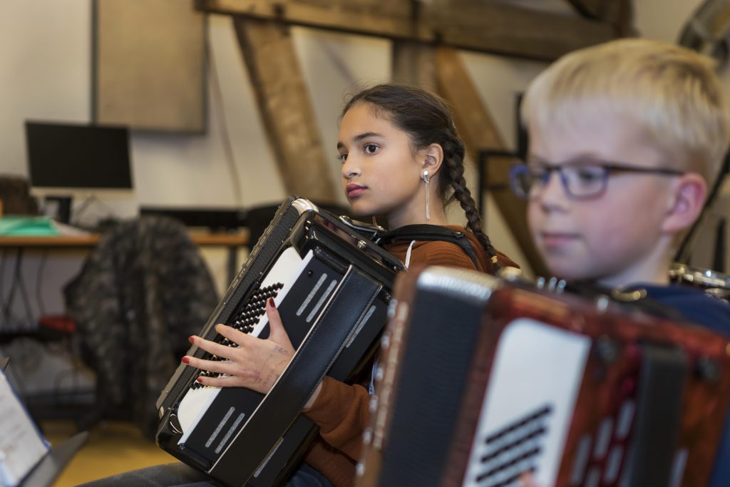 Meisje speelt accordeon tijdens een muziekles, op de voorgrond een jongetje met een accordeon.