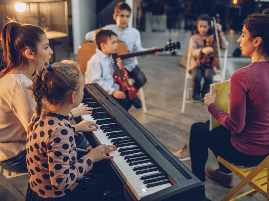 Een groep kinderen volgt samen muziekles. Op de voorgrond bespeelt een kind een digitaal pianotoetsenbord, terwijl anderen met een viool en gitaar klaarzitten. Een docent begeleidt de les vanuit een kringopstelling in een lichte, warme binnenruimte.