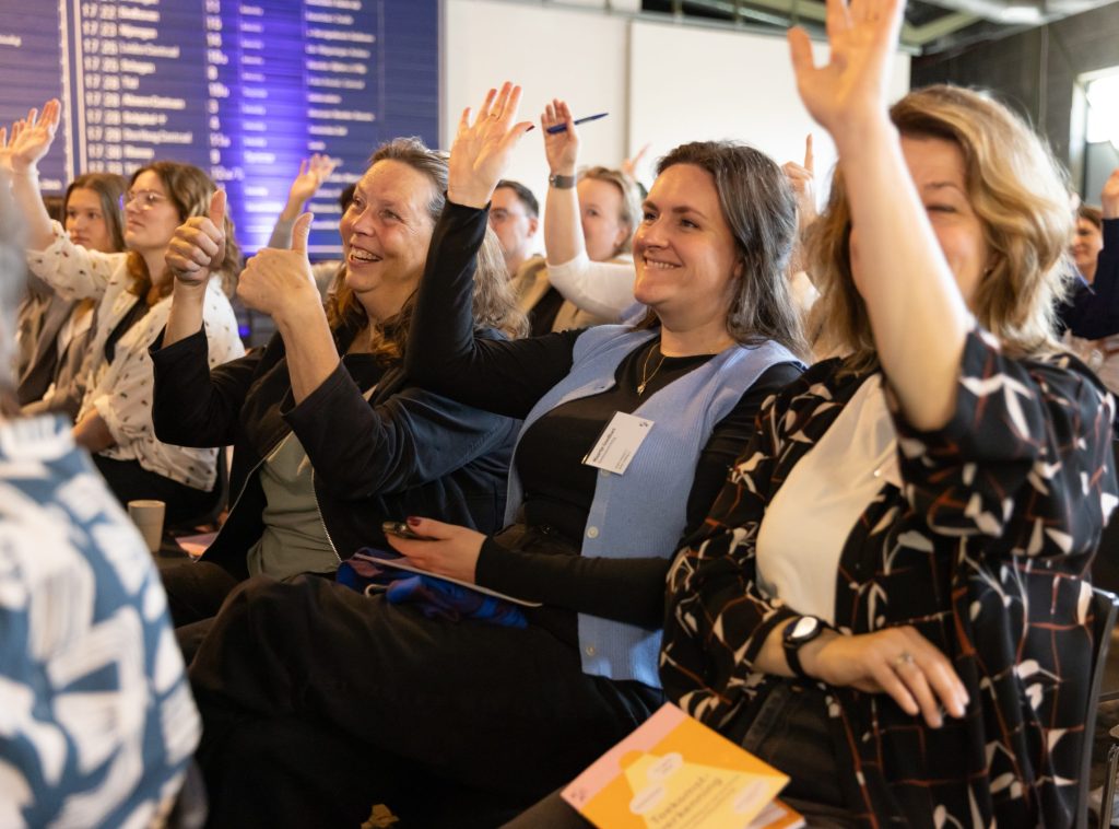 Deelnemers steken hun hand op tijdens een interactieve sessie op de conferentie.