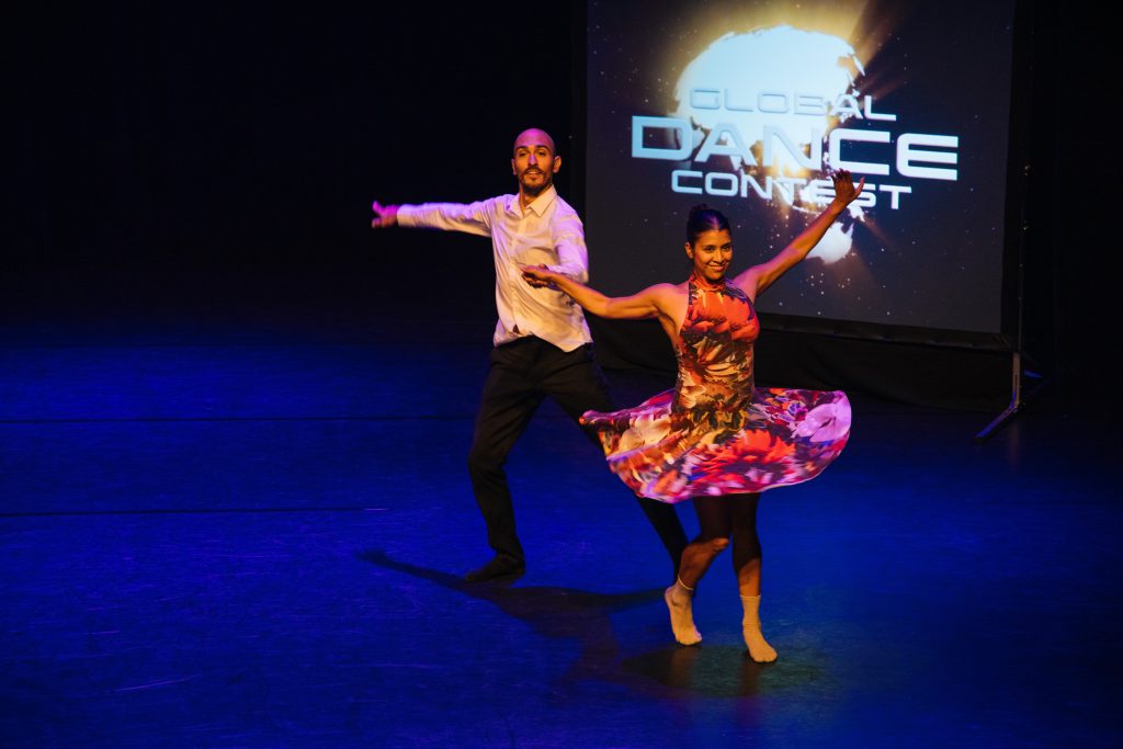 En man en een vrouw dansen op een podium met op de achtergrond een scherm met de tekst 'Global Dance Contest'.