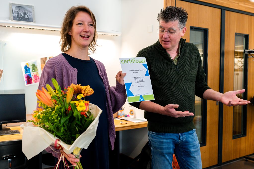 Mijke Rummens met in haar handen bloemen en het certificaat van de trainingreeks. Ronald Kox staat naast haar.