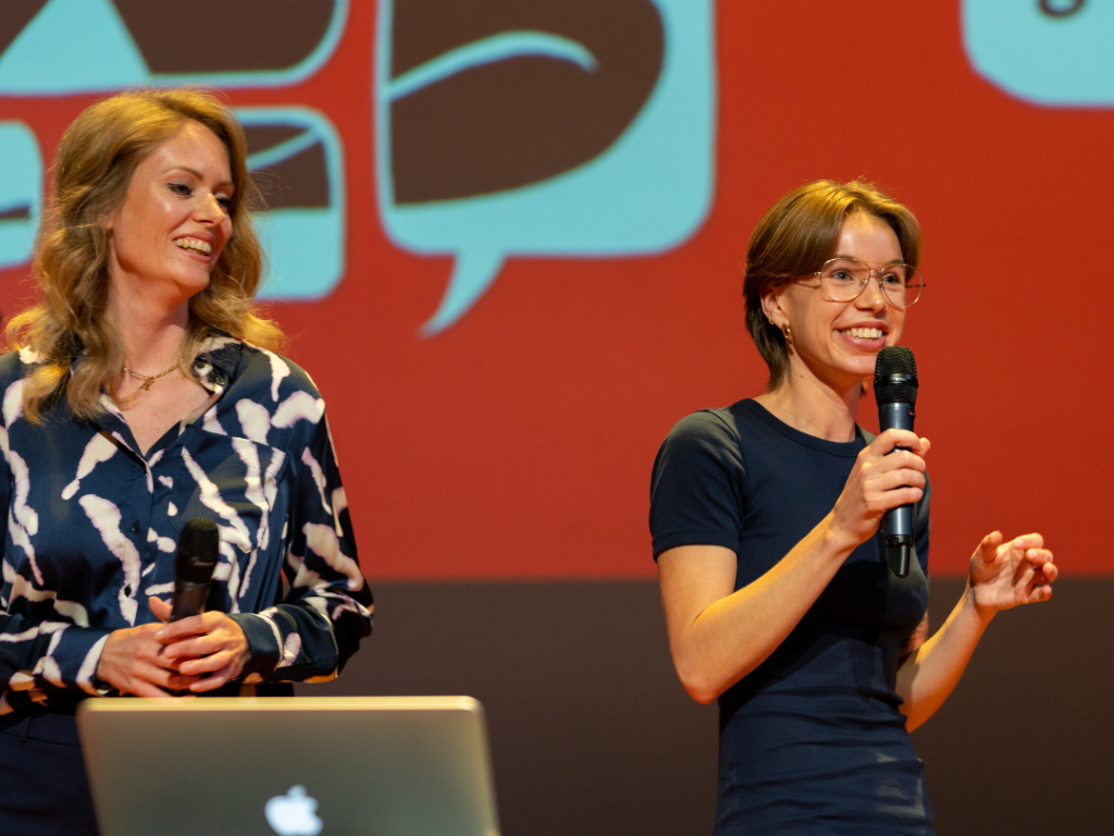 twee sprekers kijken richting het publiek en lachen