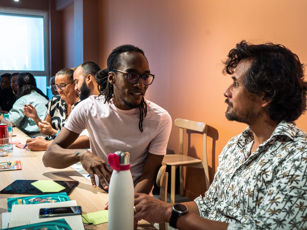 Deelnemers zitten aan een lange tafel en zijn met elkaar in gesprek tijdens een bijeenkomst. Op tafel liggen notitieblokken, telefoons en andere materialen.