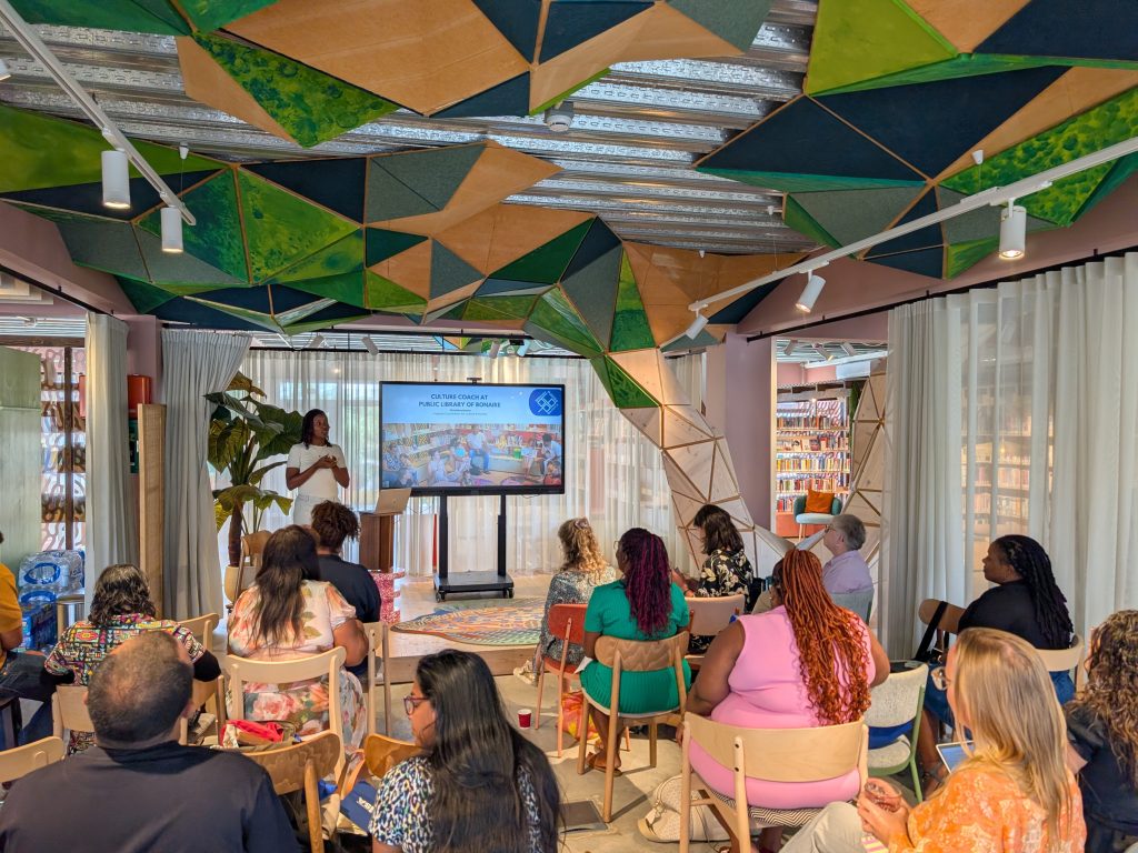 Een spreker geeft een presentatie aan een groep deelnemers in een kleurrijke ruimte. Op een groot scherm is een presentatie over cultuurcoaching te zien.