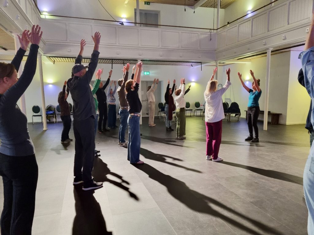 Een workshop in een danszaal waarbij meerdere mensen met beide armen omhoog staan.